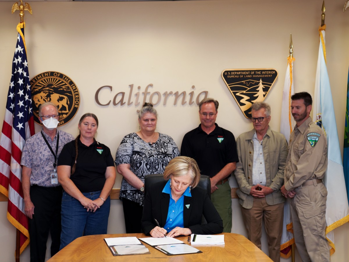 BLM California State Director Karen Mouritsen (in center sitting down) signing the Decision Record for the Statewide Wildland-Urban Interface Fuels Treatment Programmatic Environmental Assessment, with six staff members and/or partners (in back) who assisted with the project. The American Flag, state of California Flag and DOI and BLM agency flags are also displayed in the background.